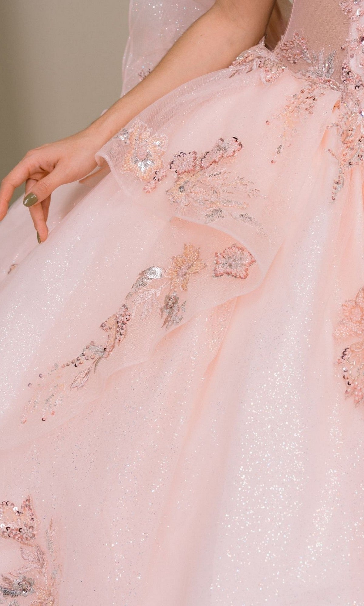 Wearing the Quinceanera Dress 8540JQ By Cinderella Couture, the person is adorned in a blush pink glitter tulle ball gown embellished with sparkling sequins and delicate floral embroidery, their arm elegantly extended at their side.